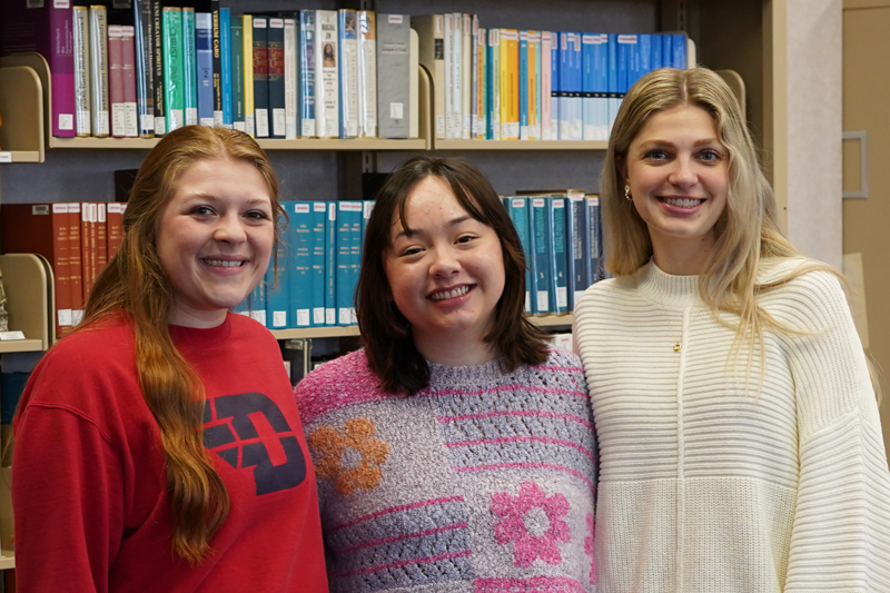 Three college students standing together and smiling in front of a wall of books.