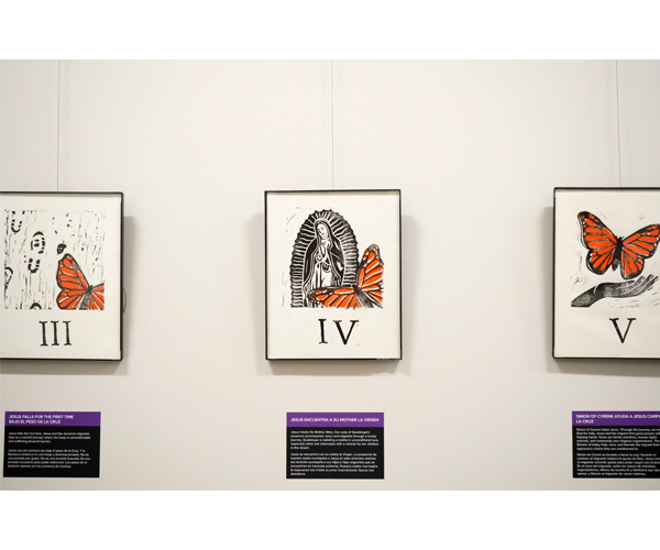 Three prints on white paper hang in thin black frames on a white wall. The prints use black and orange ink to depict Stations of the Cross with a monarch butterfly rather than Jesus Christ. Underneath each print is a black label with a purple strip across the top with white text. 
