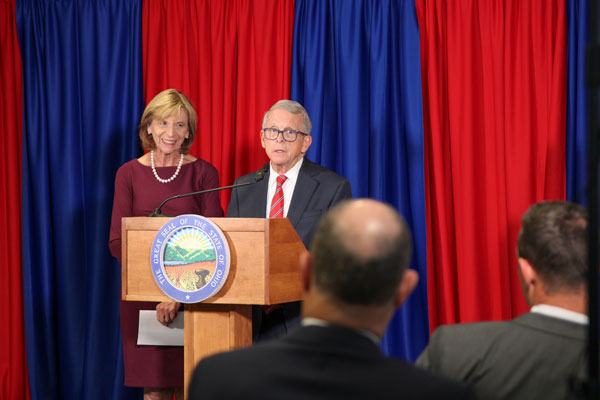 Gov. Mike DeWine at a podium with the Ohio seal on the front, in front of blue and red drapery.