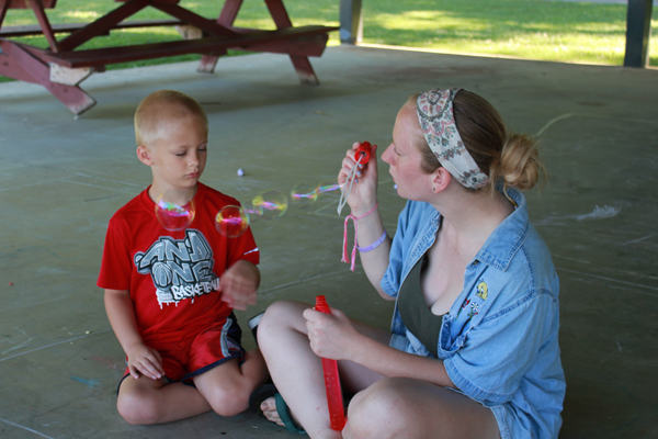 Woman sitting cross-legged on the ground blowing bubbles to a young boy in a red shirt.