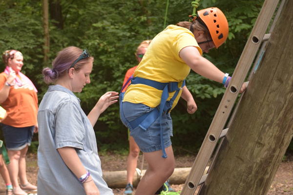 A woman is helping a young person in a yellow shirt climb up a ladder. The young person is wearing a harness for a climbing wall.