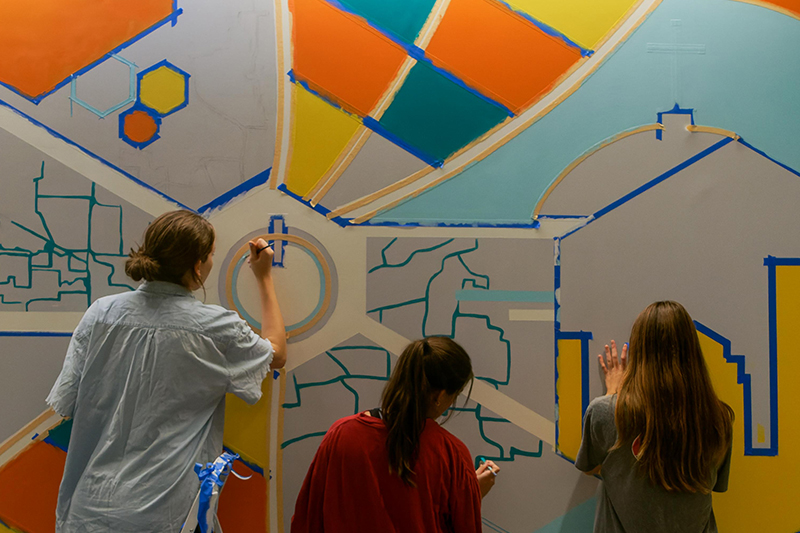 From left to right, Claire, Gracie and Mira, all with their backs turned to the camera as they are painting the mural, which is still in progress.