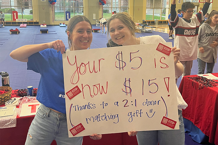 Two students, Annie Sherman on the right, holding up a sign during One Day, One Dayton that reads: "Your $5 is now $15! Thanks to a 2:1 donor matching gift"