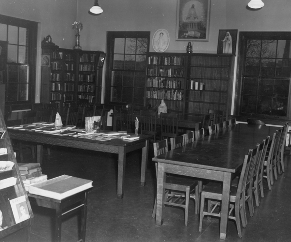 Black and white photograph of reading room, ca. 1950, with long wooden tables surrounded by chairs in the foreground. The back wall is lined with bookcases. The framed painting hangs above a middle bookcase.