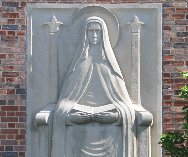 Gray in color, this bas relief is of Mary looking straight ahead with an open book on her lap. It the rectangular sculpture is surrounded by red brick of the building.