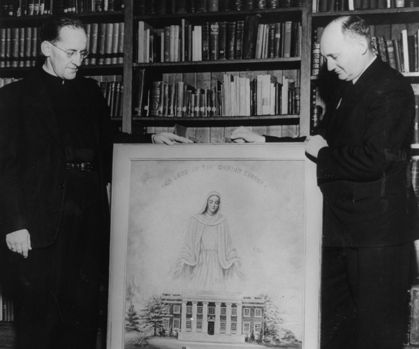 Black and white photograph of two priests in black attire with white collars standing in front of a bookcase and on each side of the painting and holding it up for the camera. 