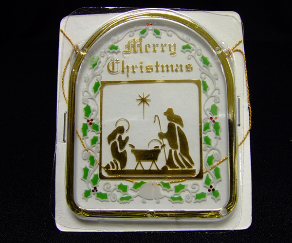 A plastic Christmas ornament made to look like glass with a gold painted frame. “Merry Christmas” is printed at the top center of the ornament, and below it is a gold foil image of the Nativity featuring Mary kneeling on the left, Christ’s manger in the middle and Joseph standing and looking down at them on the right. The image is surrounded by a foil border. A border of holly and berries painted in green and red appears between the gold frame and the gold border.