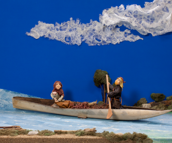 This handmade depiction of the Flight to Egypt is 3-dimensional. Dolls representing the Holy Family are in a canoe made of a natural material. Mary is dressed in brown and holds the Infant Jesus in her arms while sitting in the canoe. Joseph, dressed in dark gray, seems to be paddling the canoe from a sitting position. The canoe is placed on a hand-painted river. The banks are adorned with moss and small rocks. The sky is a very bright, even blue with one long fuzzy cloud.