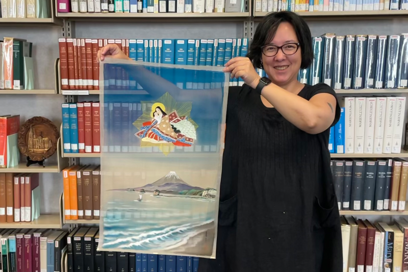 Hsuan Tsen smiles as she stands in front of a large bookcase holding a piece of artwork to the side. The artwork is on a transparent fabric and features Our Lady of Japan holding the Infant Jesus. Delicate lines of gold radiate out, forming a star behind them. They are floating in the sky above a snow-capped Mount Fuji and a body of water.