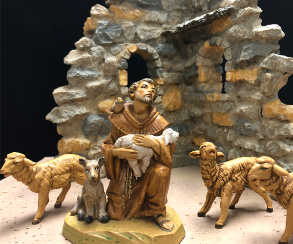 Photo of a plastic figure of a man in a brown robe who is kneeling and gazing slightly upward. He has a brown bird on his shoulder and holds a white lamb, and a dog sits by his side. Three plastic sheep stand nearby, all in front of a stone wall made of plastic.