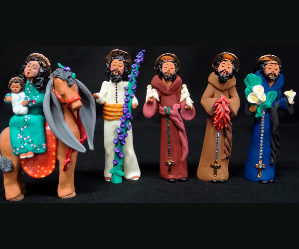  A photo of five clay figures with golden halos standing in a row with their eyes closed and mouths open in song or speech. A woman in a turquoise dress, representing the Blessed Virgin Mary, holds an infant in white clothing, representing Jesus, as she sits upon a brown and gray donkey with long, exaggerated ears. A man in white shirt and pants with an orange belt holds a tall, green staff with purple flowers. Three men in robes, each with a rosary hanging from his waist, stand next in line. The man in a burgundy robe with white birds resting on his hands represents St. Francis.