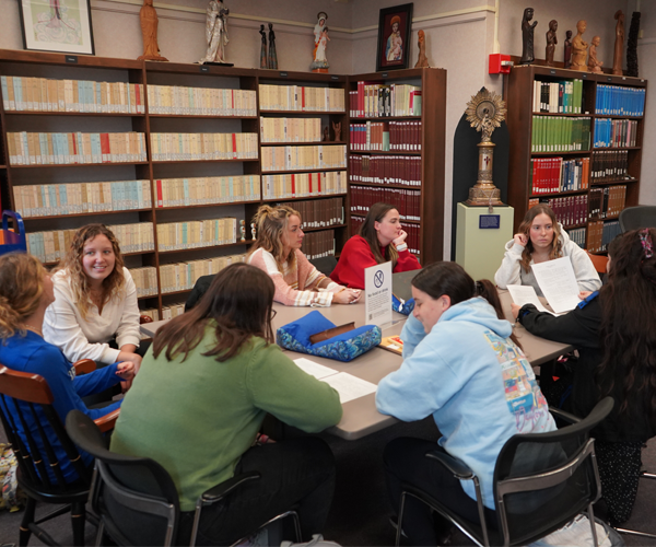 Photo of eight teacher education students at a table in the Marian Library reading room discussing the Marian Library collection items in front of them.  