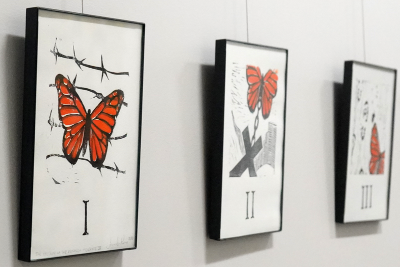 The first three of fifteen prints on white paper hang in thin black frames on a white wall. The prints use black and orange ink to depict Stations of the Cross with a monarch butterfly rather than Jesus Christ. Underneath each print is a black label with a purple strip across the top with white text. 