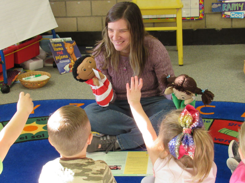 In a classroom setting, an ECHO counselor sits on carpet using two puppets to demonstrate positive emotional skills