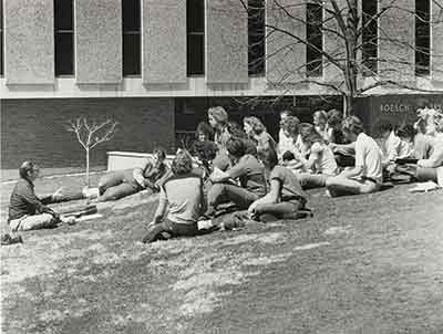 Professor teaches group os students sitting outdoors on the lawn.
